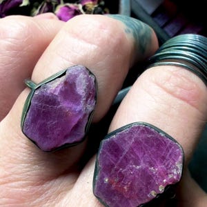 May include: Two rings featuring large, raw, deep magenta-colored gemstones set in dark, oxidized metal bands. The stones have irregular, faceted surfaces with a natural, unpolished texture, held securely by thin, dark metal settings.