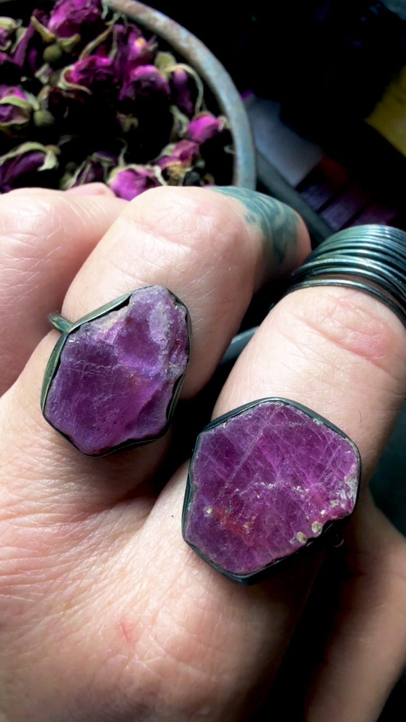 May include: Two rings featuring large, raw, deep magenta-colored gemstones set in dark, oxidized metal bands. The stones have irregular, faceted surfaces with a natural, unpolished texture, held securely by thin, dark metal settings.