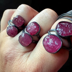 以下が含まれることがあります： シルバー色の金属にセットされた、大きく不規則な形の濃いピンク色の宝石を特徴とするいくつかのリングのクローズアップ。宝石は粗く自然な質感です。リングは暗い背景に対して手で表示されます。