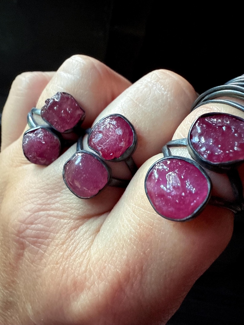 以下が含まれることがあります： シルバー色の金属にセットされた、大きく不規則な形の濃いピンク色の宝石を特徴とするいくつかのリングのクローズアップ。宝石は粗く自然な質感です。リングは暗い背景に対して手で表示されます。