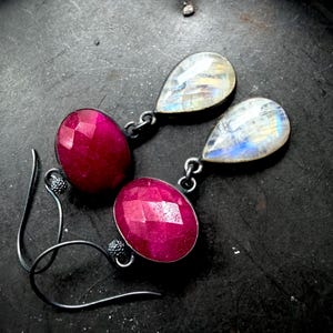 May include: A pair of handcrafted earrings featuring faceted, oval-cut, deep pink gemstones paired with teardrop-shaped, iridescent moonstones. The earrings have a dark metal setting and ear wires, showcasing a unique contrast of colors and textures, perfect for adding a touch of elegance.