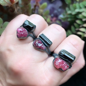 May include: Three silver rings with black tourmaline and pink tourmaline stones. The rings are stacked on a person's finger.