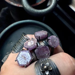 Puede incluir: Una colección de anillos de plata con piedras preciosas moradas en bruto. Uno tiene una piedra hexagonal, otros formas irregulares. También se ve un anillo floral y otro enrollado.