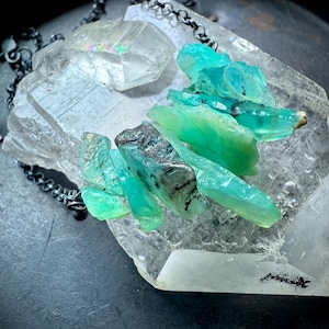 May include: A necklace featuring raw, teal-colored crystal clusters and clear quartz stones. The necklace has a dark, oxidized chain. The crystals are arranged on a large, clear quartz base, creating a natural, earthy aesthetic. The necklace is displayed on a dark surface.