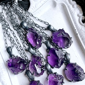 May include: A set of eight amethyst crystal pendants on silver chains. The crystals are raw and have a deep purple color. The chains are delicate and have a silver finish.