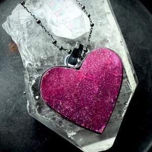 May include: A heart-shaped pendant with a deep magenta, textured surface and a dark metal rim. The pendant hangs from a thin, dark beaded chain and rests against a large, clear, multifaceted quartz crystal with visible internal fractures.