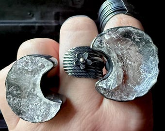 Può includere: Una mano mostra diversi anelli d'argento. Gli anelli presentano vari design, tra cui una forma a mezzaluna, una fascia strutturata con un fiore e un design a filo stratificato. Gli anelli hanno un aspetto rustico e artigianale.