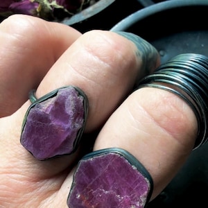 May include: Two rings featuring raw, uncut, deep magenta-colored gemstones set in dark, oxidized metal bands. The stones have irregular, jagged edges and a natural, unpolished texture. One ring is worn on the index finger and one on the middle finger.