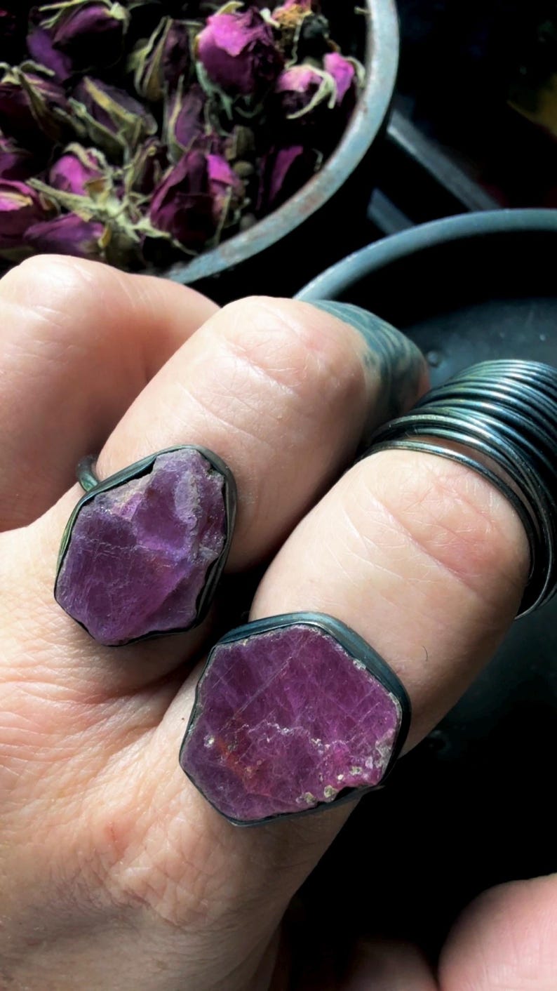 May include: Two rings featuring raw, uncut, deep magenta-colored gemstones set in dark, oxidized metal bands. The stones have irregular, jagged edges and a natural, unpolished texture. One ring is worn on the index finger and one on the middle finger.