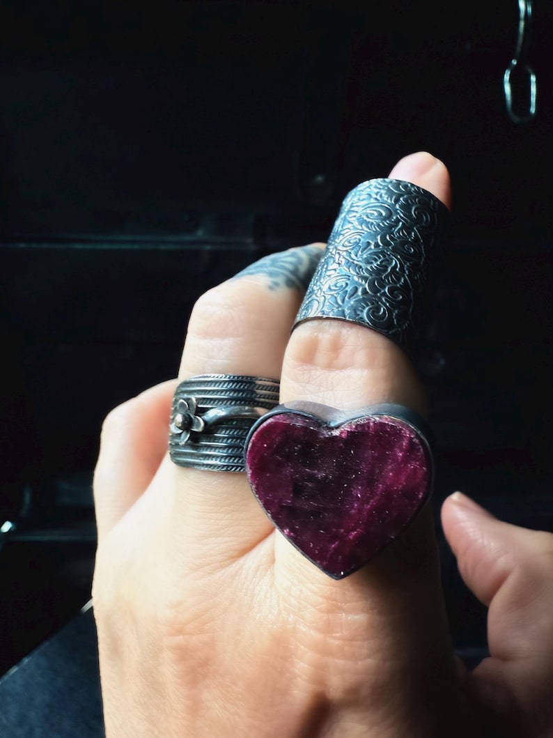 May include: A hand displays four silver rings. One ring is a long, textured band. Another is a heart-shaped ring with a deep red stone. A third ring has a floral design. The rings are set against a dark background.