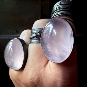 May include: A hand wearing several rings, including two large oval-shaped rings with pale pink stones and silver bands. Other rings include a floral design and a stack of thin silver bands. The background is dark.