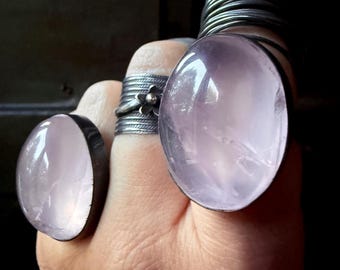 Può includere: Primo piano di una mano con diversi anelli. Due anelli importanti presentano grandi pietre preziose ovali rosa pallido incastonate in argento. Altri anelli includono un motivo floreale e una pila di sottili fasce d'argento. Lo sfondo è scuro.