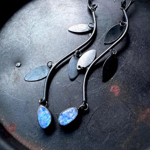 May include: A pair of long, silver earrings with a leaf design. Each earring features a curved stem with several small, flat leaves and a teardrop-shaped, blue opal stone at the bottom. The earrings are set against a dark, textured background.