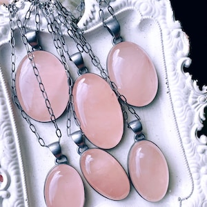 May include: Six oval-shaped pink rose quartz pendants on silver chains. The pendants are arranged in a white decorative tray.