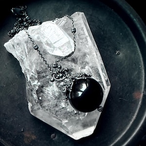 May include: A black gemstone pendant necklace with an ornate silver-tone setting and chain. The pendant is a large, round, polished black stone. The necklace rests on a clear quartz crystal, set against a dark background.