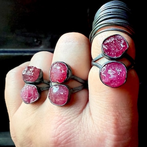 以下が含まれることがあります： 手には、それぞれ生の鮮やかなピンク色の宝石が付いた、いくつかのシルバーリングがはめられています。 リングはシンプルなデザインで、石が際立っています。 背景は暗く、ジュエリーが強調されています。