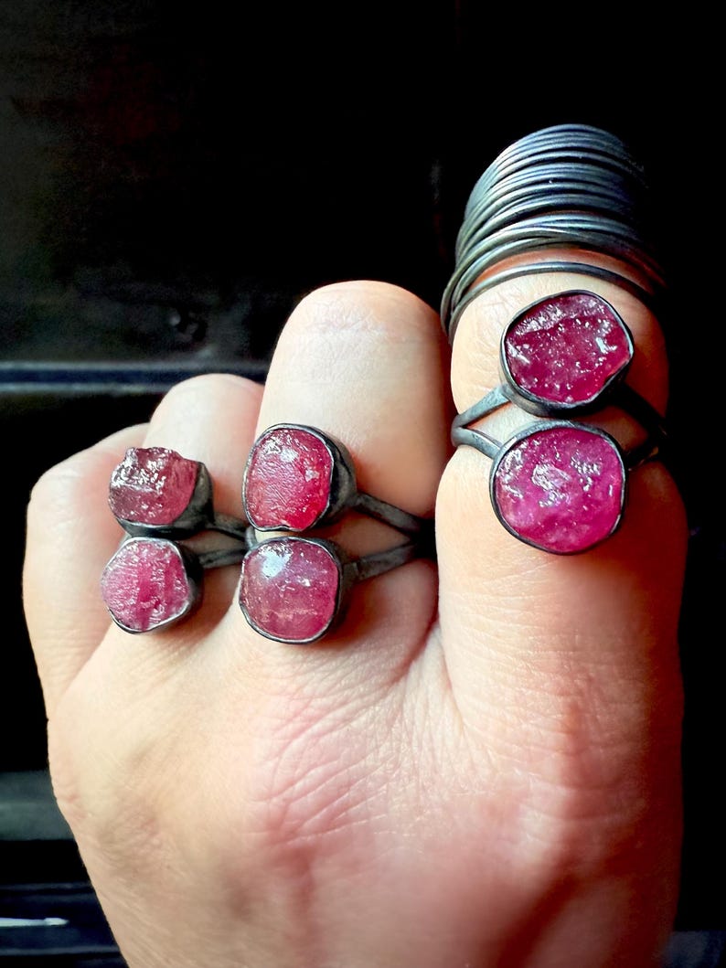 以下が含まれることがあります： 手には、それぞれ生の鮮やかなピンク色の宝石が付いた、いくつかのシルバーリングがはめられています。 リングはシンプルなデザインで、石が際立っています。 背景は暗く、ジュエリーが強調されています。