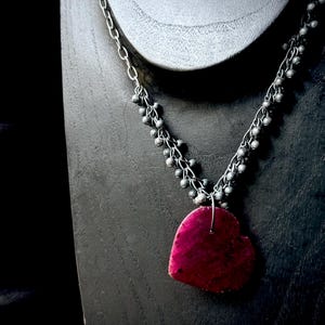 May include: A silver necklace with a heart-shaped, deep pink pendant. The necklace features a chain with small, round beads and a larger, irregular heart pendant. The jewelry is displayed on a dark grey mannequin.
