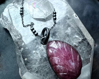 May include: A dark silver necklace with a pendant featuring a carved, oval-shaped, deep pink gemstone. The pendant is suspended from a delicate chain with small, dark beads. The necklace rests on a large, clear crystal.