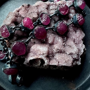 May include: A necklace featuring raw, deep pink gemstones set in dark metal links. The necklace is displayed on a rough, textured, light purple mineral formation, creating a contrast of textures and colors.