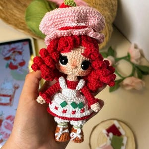 Pode incluir: Uma boneca de croch&ecirc; com cabelo ruivo, um chap&eacute;u rosa, um vestido branco com detalhes verdes e vermelhos e sapatos marrons. A boneca est&aacute; segurando um pequeno objeto verde na m&atilde;o.