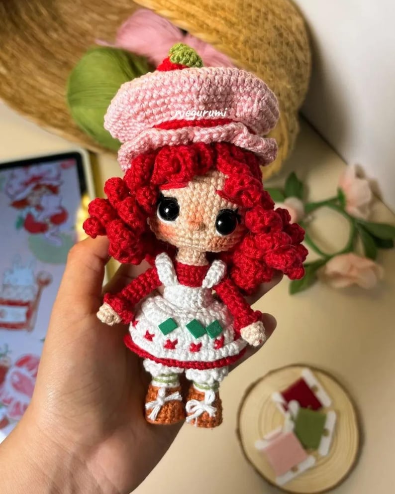 Pode incluir: Uma boneca de croch&ecirc; com cabelo ruivo, um chap&eacute;u rosa, um vestido branco com detalhes verdes e vermelhos e sapatos marrons. A boneca est&aacute; segurando um pequeno objeto verde na m&atilde;o.