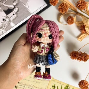 May include: A handmade crochet doll with pink hair, wearing a gray skirt, a beige jacket, and burgundy shoes. The doll is holding a blue bag. The doll's name, "Niegurumi", is written on the image.