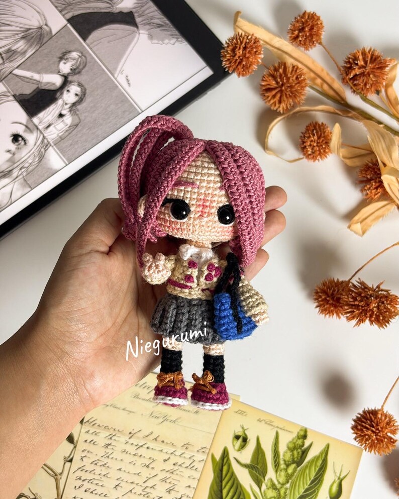 May include: A handmade crochet doll with pink hair, wearing a gray skirt, a beige jacket, and burgundy shoes. The doll is holding a blue bag. The doll's name, "Niegurumi", is written on the image.