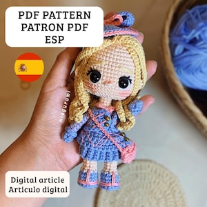Puede incluir: Muñeca de ganchillo con cabello rubio, vestida con un vestido azul y un bolso rosa. La muñeca tiene grandes ojos negros y mejillas rosadas. El texto en la imagen dice "PDF PATTERN PATRON PDF ESP" y "Digital article Articulo digital".