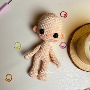 May include: A crocheted amigurumi doll in a light peach color. The doll has large black eyes, small ears, and a blush on its cheeks. The doll is standing with its arms and legs extended. The word "Niegurumi" is written on the doll's body.