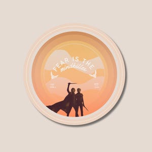 May include: Round sticker with a sunset-colored background and the text "FEAR IS the mindkiller." Two silhouetted figures stand with weapons. The sticker has a cream-colored border and the text "TND DUNE II" and "MRK 2024".