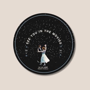 Puede incluir: Una pegatina redonda negra con el texto blanco "SEE YOU IN THE MOVIES". Presenta una pareja bailando, el título de la película "LA LA LAND" y el nombre del director Damien Chazelle. También incluye las fechas "SEB 20" y "MIA 16".