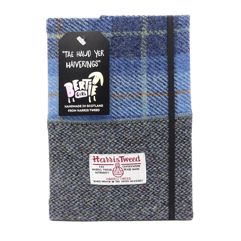 May include: A blue and gray plaid notebook cover with a black elastic closure. The cover is made from Harris Tweed and features a label that reads "Tae Haud Yer Haiverings" and a small sheep illustration. The label also states "Handmade in Scotland from Harris Tweed".