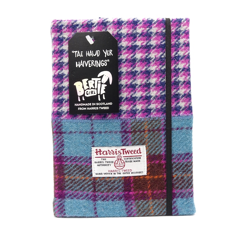May include: A pink and blue plaid notebook with a black elastic closure. The cover is made of Harris Tweed fabric and features a black tag with the text "TAE HAUD YER HAIVERINGS" and a small sheep illustration. The tag also includes the text "Bertie Girl Handmade in Scotland from Harris Tweed".