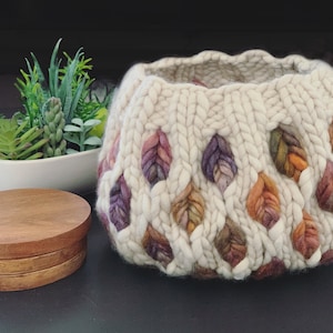 May include: A cream-colored, hand-knitted basket with leaf-shaped accents in shades of purple, orange, and brown. The basket is made of thick yarn and has a rounded shape. A stack of wooden coasters and a small succulent plant in a white bowl are also visible.