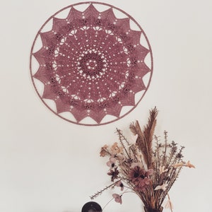 May include: A large, dusty rose crochet mandala wall hanging with a brown metal pipe and a vase of dried flowers with pink and brown blooms.