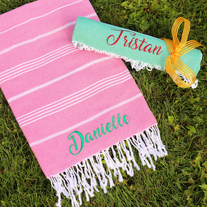May include: A pink and white striped beach towel with white fringe. The towel has the name "Danielle" embroidered on it in green. A rolled up green towel with the name "Tristan" embroidered on it in red is lying on top of the pink towel.