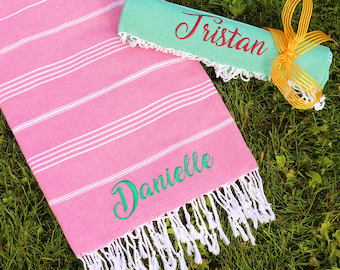 Op de afbeelding: Een roze-wit gestreepte strandhanddoek met witte franjes. De handdoek heeft de naam "Danielle" geborduurd in het groen. Een opgerolde groene handdoek met de naam "Tristan" geborduurd in het rood ligt op de roze handdoek.