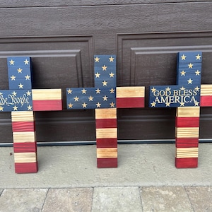 May include: Three wooden crosses, each with a design inspired by the American flag. They feature blue sections with gold stars and red and white stripes. One cross has the text "We the People," and another reads "God Bless America."