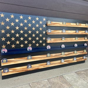 Puede incluir: Expositor de madera con temática de la bandera estadounidense, con estrellas doradas sobre fondo negro. Cuenta con múltiples estantes de madera para guardar monedas conmemorativas. El diseño de la bandera incluye una franja azul y una base negra.