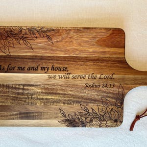 May include: A wooden chopping board with a handle and a floral design. The board has a quote engraved on it: "As for me and my house, we will serve the Lord. Joshua 24:15".