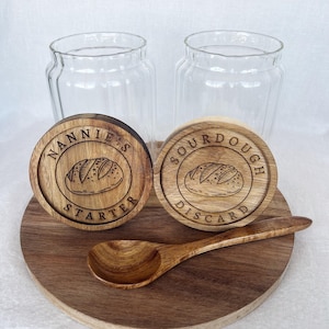 May include: Two glass jars with wooden lids, one labeled "Nannie's Starter" and the other "Sourdough Discard". Both lids feature a stylized illustration of a loaf of bread. A wooden spoon is also included.
