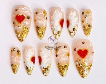 May include: Set of ten false nails with a white and gold glitter design. Each nail features a red heart and a white flower design. The nails are shaped like a stiletto.