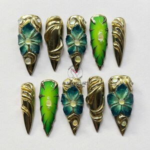 Op de afbeelding: Een set van tien goudkleurige, puntige valse nagels. Sommige nagels hebben een groen verloopontwerp, terwijl andere een blauwgroene bloem met een centrale edelsteen hebben. De nagels zijn versierd met gouden accenten en kleine edelstenen.