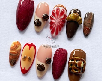 May include: A collection of ten almond-shaped acrylic nails with various designs. The nails feature colors like burgundy, nude, red, and brown, with patterns including coffee beans, fruit slices, and textured surfaces. The text "ALISA NAILS ART LOVE LIFE DESIGN" is visible.