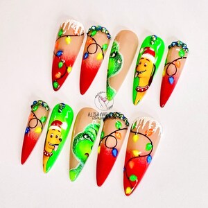 May include: Set of ten long, pointed, false nails with Christmas-themed designs. The nails are painted with a red, white and green gradient, and feature images of the Grinch, a dog wearing a Santa hat, and Christmas lights.