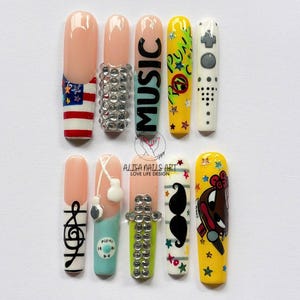 May include: A collection of ten artificial nails with various designs. Some nails feature the American flag, rhinestones, the word "MUSIC", music notes, and cartoon characters. The nails are painted in a variety of colors, including pink, yellow, and white.