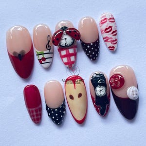 May include: A set of ten artificial nails with various designs. The nails feature a combination of red, black, white, and beige colors. Designs include ladybugs, musical notes, polka dots, and lip prints. Some nails have 3D embellishments.