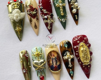 May include: A collection of ten long, almond-shaped artificial nails. Each nail features intricate designs, including crosses, hearts, and religious imagery, adorned with rhinestones, pearls, and gold accents. The nails are in various colors, including gold, red, green, and nude.