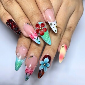 May include: A set of long, pointed acrylic nails with various designs. The nails feature a range of colors, including red, pink, blue, and white, with floral and abstract patterns. Some nails have 3D embellishments, such as flowers and rhinestones.